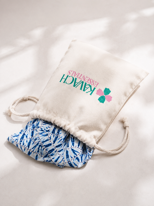 An off-white cotton drawstring pouch with 'KAVACH ESSENTIALS' printed on the front, containing a blue and white patterned fabric item, set against a softly lit white background with dappled shadows.