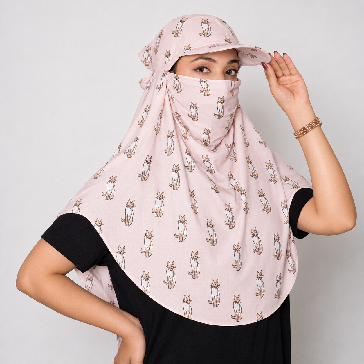 A woman wearing a light pink sun protection headscarf with a repeating cat print, paired with a matching sun hat and a black t-shirt, against a plain light gray background.