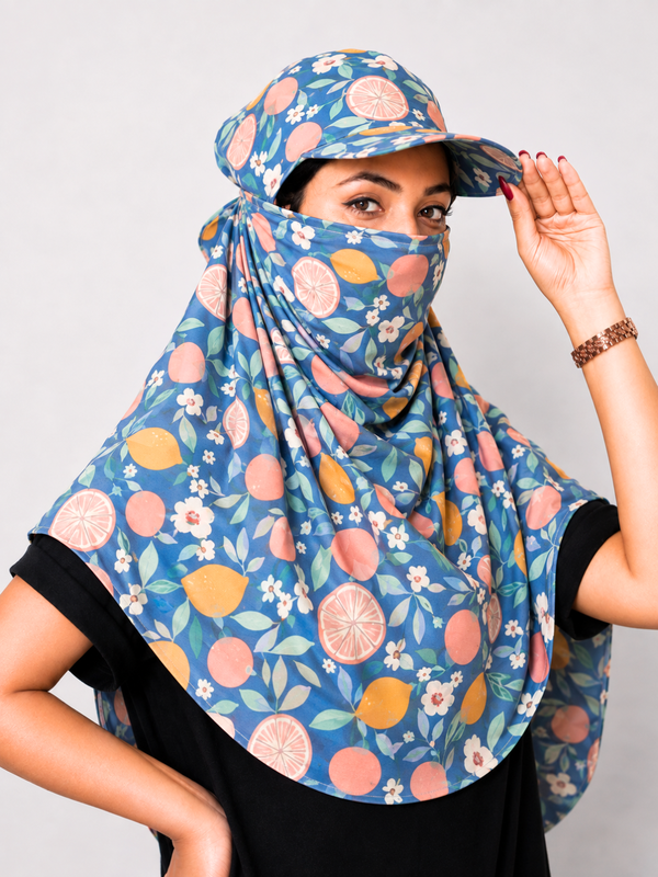 A woman wearing a blue sun protection set featuring a matching sun hat, face mask, and neck gaiter with a citrus and floral print, posed against a light gray background.