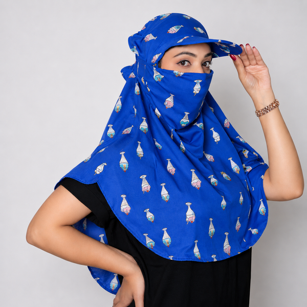 A woman wearing a royal blue hijab with a repeating pattern of colorful perfume bottles, paired with a matching blue sun hat. She is posing against a solid light gray background.