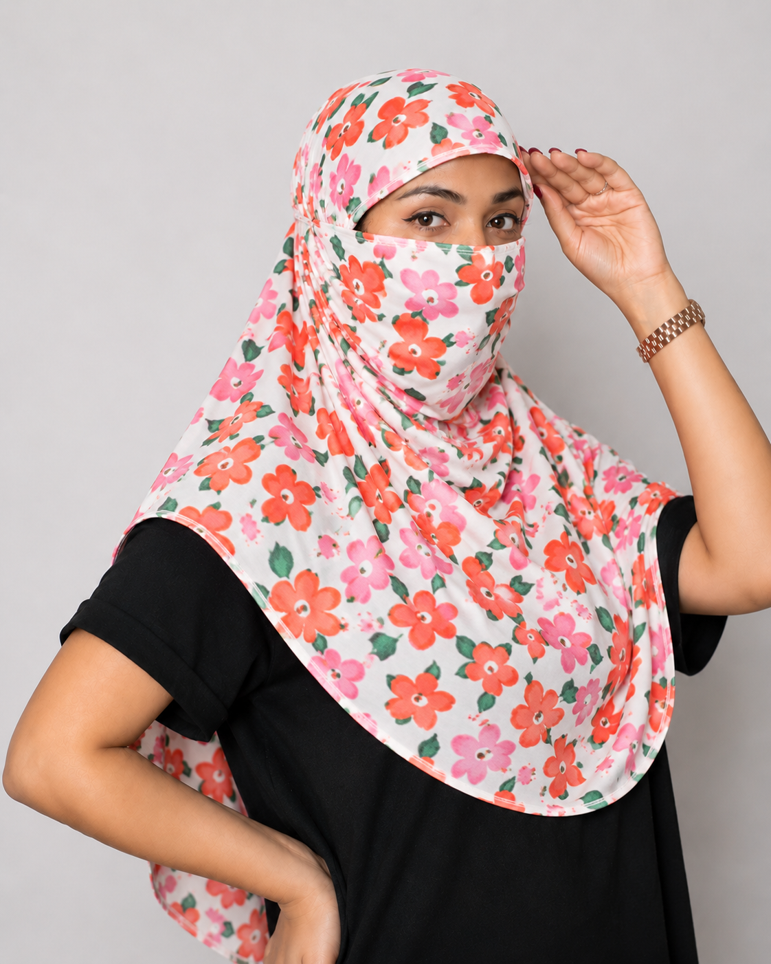A woman wearing a white face scarf with a vibrant pink, red, and green floral print, styled over a black t-shirt against a solid gray studio background.