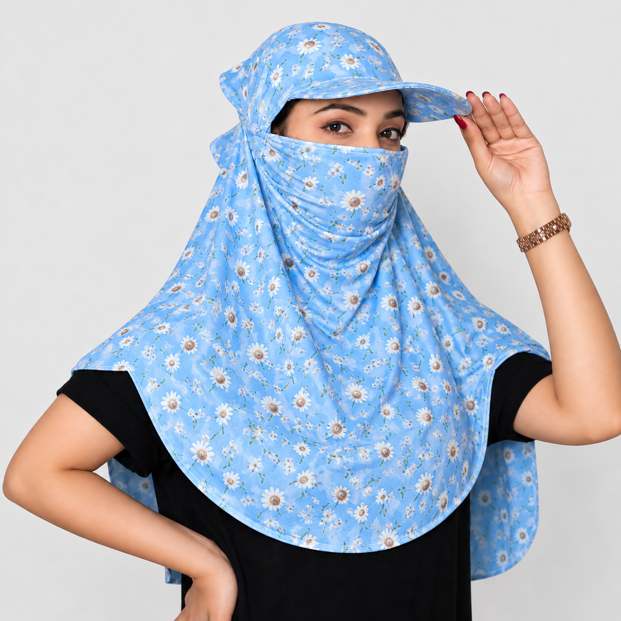 A woman wearing a light blue sun protection headscarf with a daisy floral print, featuring a built-in sun hat and neck gaiter, over a black t-shirt against a light gray background.