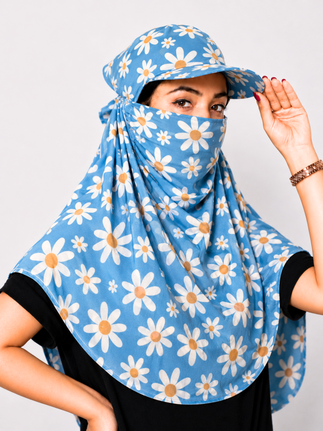 A woman wearing a light blue sun protection headscarf with a white daisy pattern, featuring a matching visor and neck gaiter, set against a plain light gray background.