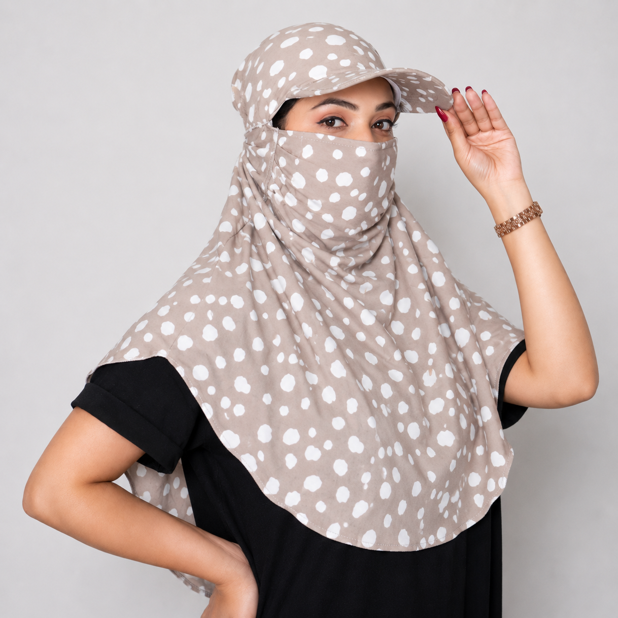 A woman wearing a beige and white polka dot hijab with a matching sun cap, posing against a solid gray studio background.
