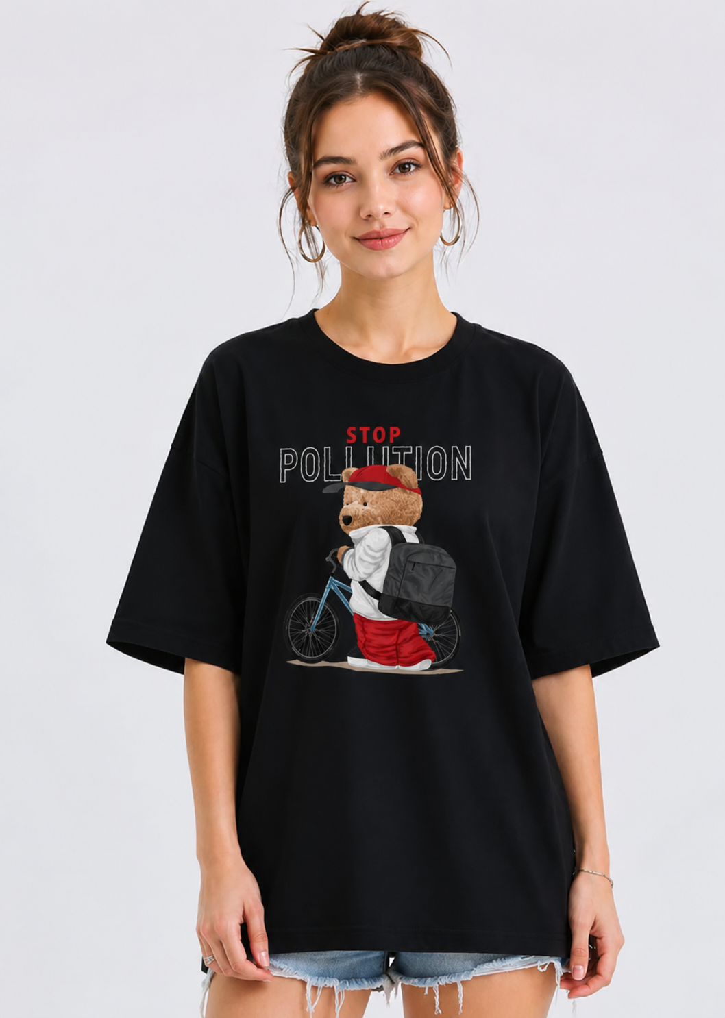 A woman wearing a black oversized graphic t-shirt with a cartoon bear riding a bicycle and the words 'STOP POLLUTION' printed on the front, paired with frayed denim shorts, against a light gray studio background.