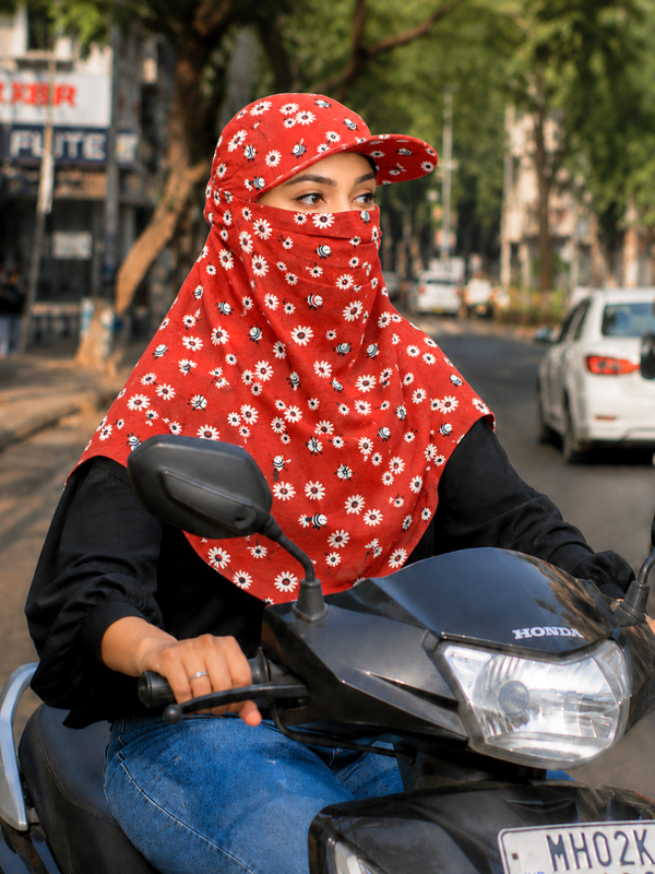 A woman wearing a red floral face scarf and matching cap rides a black two wheeler on a city street.