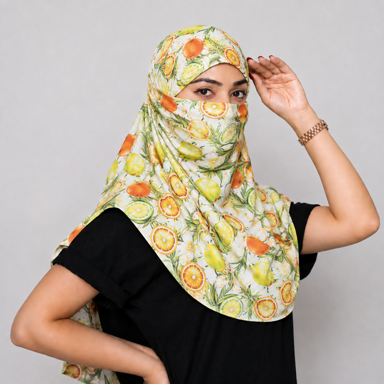 A woman wearing a printed face scarf with a citrus and fruit pattern, paired with a black t-shirt, posing against a solid gray studio background.