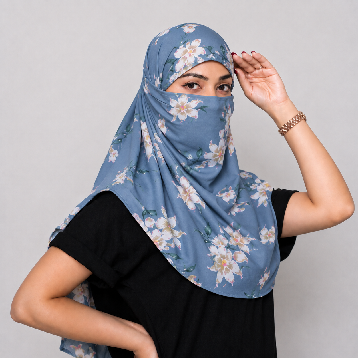 A woman wearing a dusty blue floral sun protection face scarf and a black t-shirt, posing against a neutral gray background.