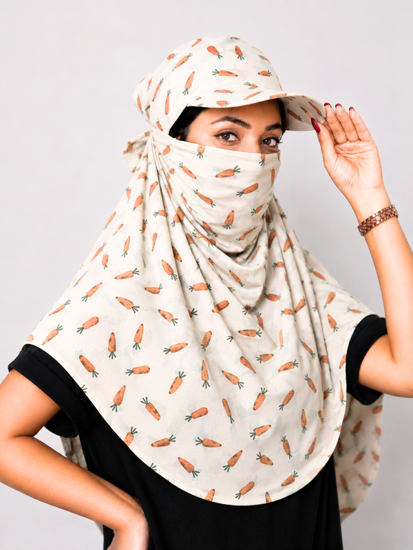 A woman wearing a beige sun protection headscarf with a repeating orange carrot print, paired with a matching beige sun hat and a black t-shirt, posing against a light gray background.