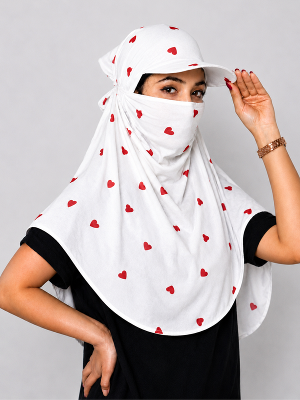 A woman wearing a white sun protection headscarf with a matching cap and face mask, decorated with small red hearts, against a solid gray background.