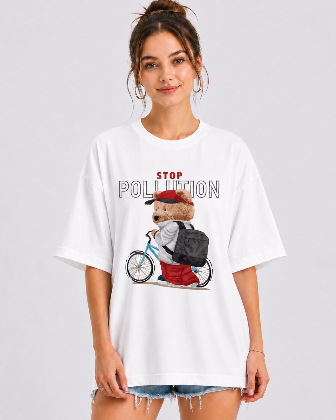 A woman wearing an oversized white graphic t-shirt with a cartoon bear riding a bicycle and the words 'STOP POLLUTION' printed on the front, paired with denim shorts, against a light gray studio background.