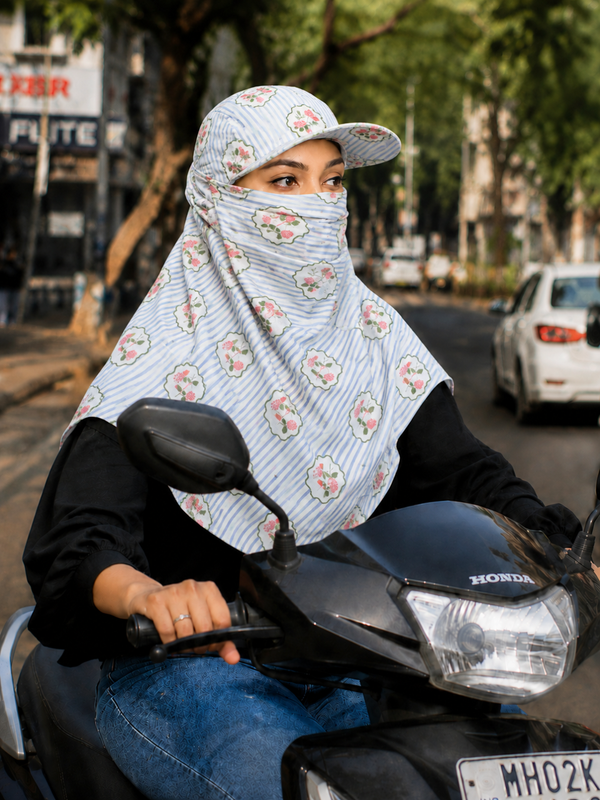 A woman riding a two wheeler on a city street, wearing a light blue and white striped sun scarf with a pink and green floral pattern, a matching cap, and a black long-sleeve top.