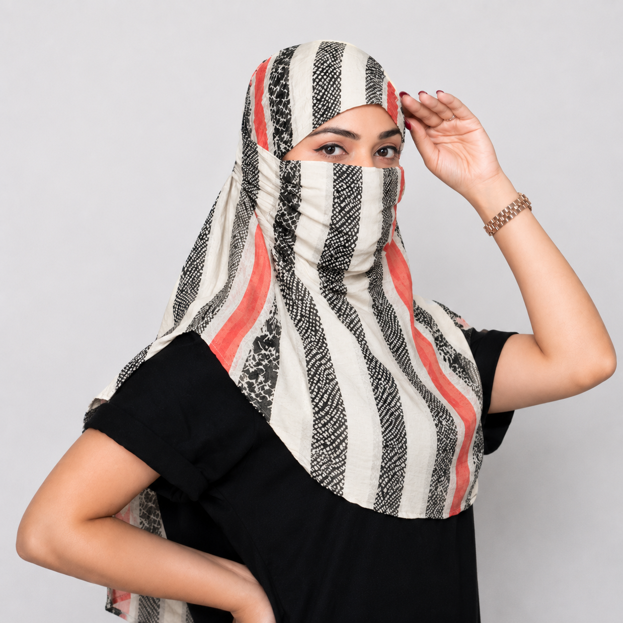 A woman wearing a patterned face scarf with vertical black, white, and coral-red stripes, paired with a black t-shirt against a light gray background.