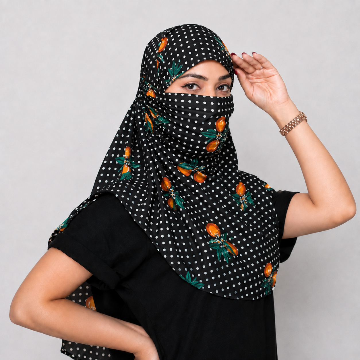 A woman wearing a black women face protection scarf with white polka dots and orange floral motifs, styled over a black top against a plain gray background.