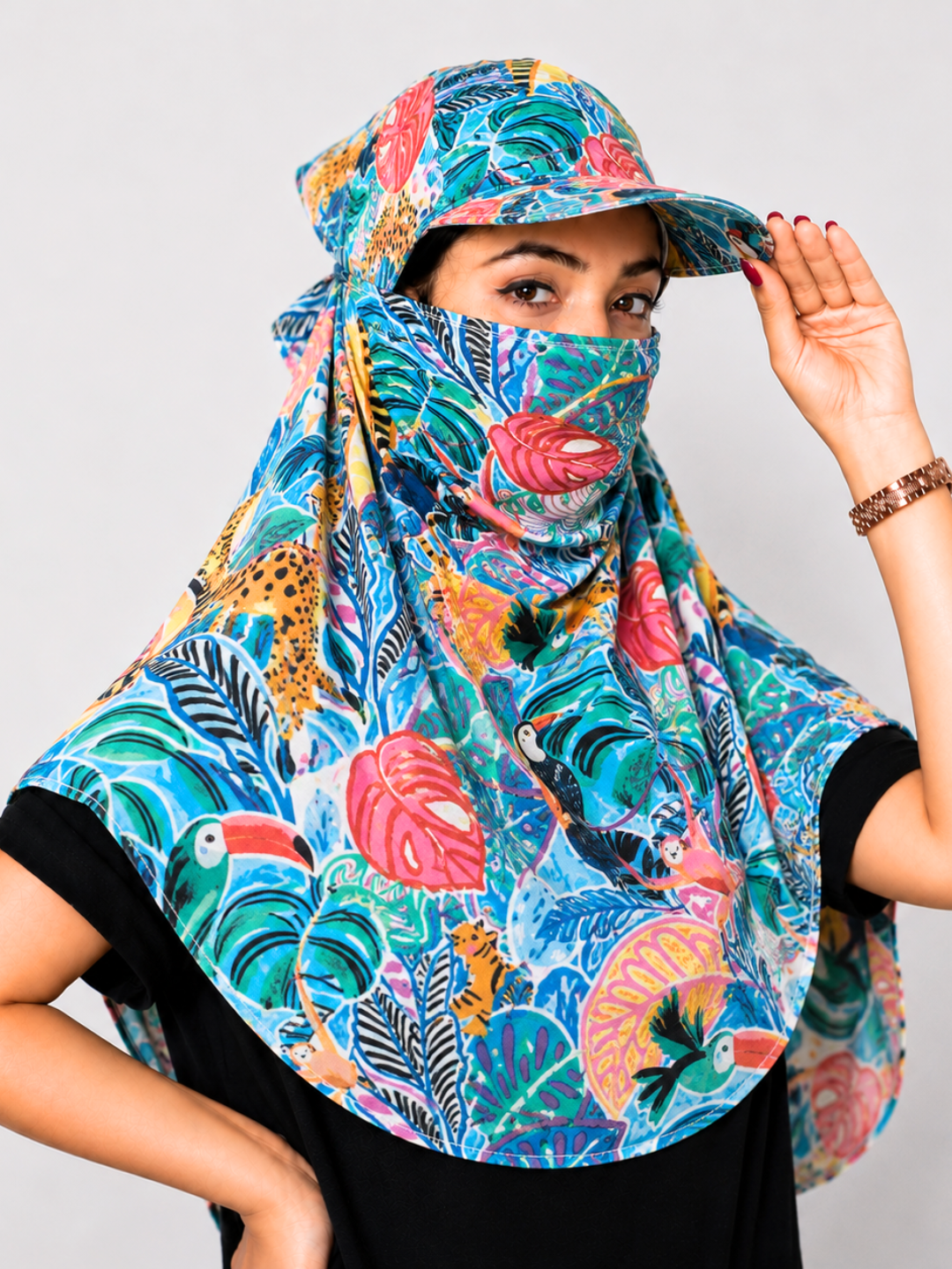 A woman wearing a vibrant tropical print sun cap and matching face covering scarf, featuring colorful toucans, leaves, and birds on a light gray background.
