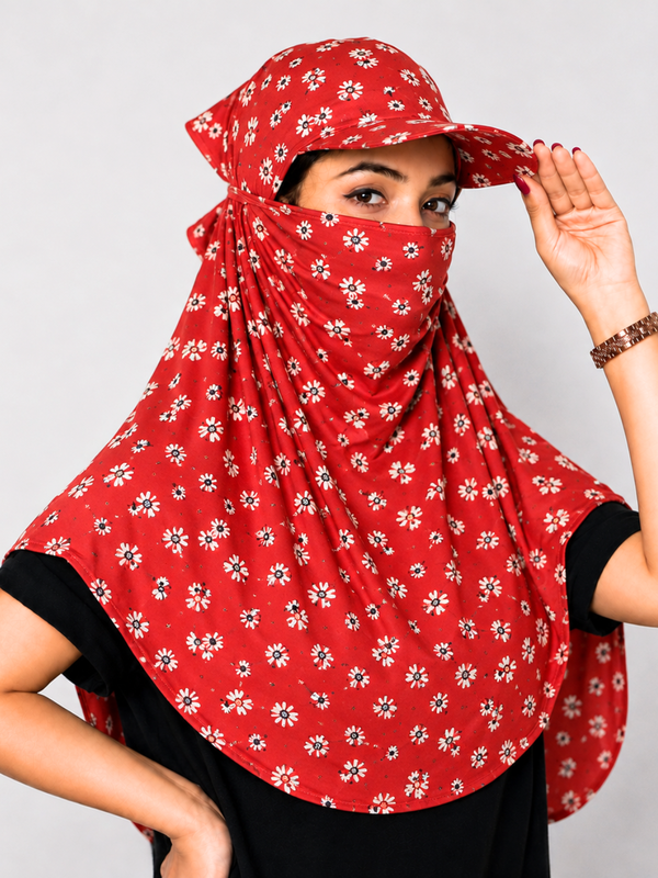 A woman wearing a red sun protection headscarf with a white and black daisy pattern, featuring a built-in sun cap and face veil, paired with a black t-shirt against a light gray background.