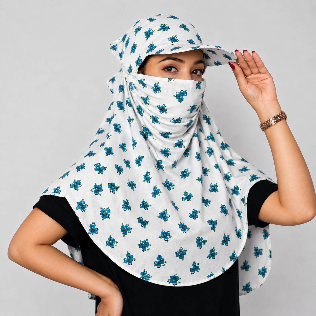 A woman wearing a white head scarf-style face veil with a blue floral pattern, paired with a matching cap and a black top, posing against a light gray background.