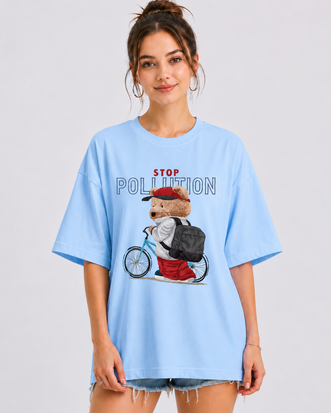 A woman modeling an oversized light blue t-shirt with a 'STOP POLLUTION' graphic featuring a cartoon bear on a bicycle. The background is a clean, neutral gray studio backdrop.
