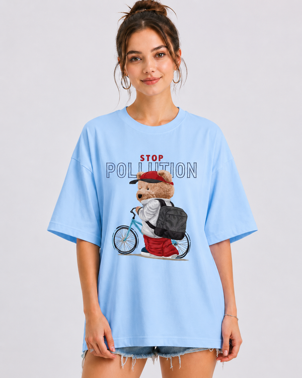 A woman modeling an oversized light blue t-shirt with a 'STOP POLLUTION' graphic featuring a cartoon bear on a bicycle. The background is a clean, neutral gray studio backdrop.